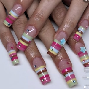 Op de afbeelding: Lange, vierkante acrylnagels met een pastelkleurig streepjesontwerp. De strepen bevatten roze, geel, blauw en bruin. Sommige nagels zijn versierd met kleine edelstenen en stervormige versieringen, wat een vleugje glans aan de manicure toevoegt.