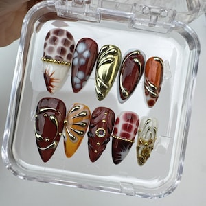 May include: A set of ten artificial nails in a clear plastic case. The nails feature various designs, including gold accents, floral patterns, and abstract shapes in shades of red, orange, and white. Some nails have a gold metallic finish.