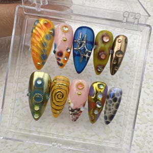 Könnte beinhalten: Ein Set mit zehn falschen Nägeln mit verschiedenen Designs. Die Nägel befinden sich in einer durchsichtigen Plastikbox. Die Designs beinhalten Spiralen, Punkte und Sterne. Die Nägel sind in verschiedenen Farben, darunter Gelb, Blau, Rosa und Braun.