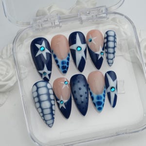 May include: A set of ten artificial nails in a clear plastic case. The nails feature a variety of designs, including navy blue with white star accents, light blue and white checkered patterns, and nude-colored tips with star details. Each nail is adorned with a small, blue gem.