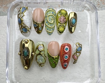 May include: A collection of ten artificial nails displayed in a clear case. The nails showcase diverse designs, including gold detailing, gemstones, and intricate patterns in silver, pink, green, and blue. The nails are oval-shaped.