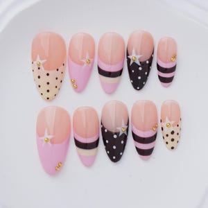 May include: A set of ten almond-shaped press-on nails featuring a nude base with various designs including pink, black, and cream stripes, polka dots, and white star accents. Each nail is embellished with small gold-toned metallic studs.