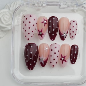 May include: A set of artificial nails in a clear plastic case. The nails feature various designs, including pink with burgundy polka dots, nude with star accents, and burgundy with white polka dots. Some nails have rhinestone embellishments.