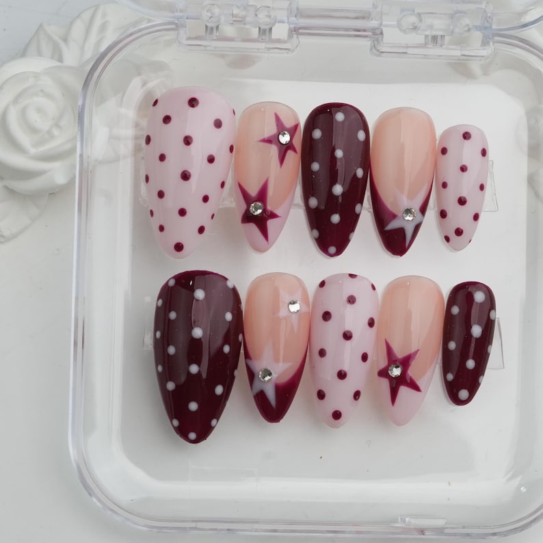 May include: A set of artificial nails in a clear plastic case. The nails feature various designs, including pink with burgundy polka dots, nude with star accents, and burgundy with white polka dots. Some nails have rhinestone embellishments.