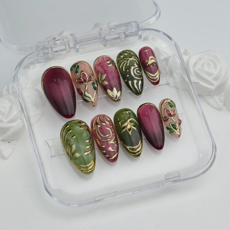 May include: A set of ten artificial nails in a clear plastic case. The nails feature a variety of designs, including solid burgundy, olive green, and pink with gold floral and swirl accents. The nails are oval-shaped.