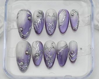 May include: A set of ten artificial nails in a clear plastic case. The nails feature a gradient design of white and purple, with silver embellishments and heart-shaped accents. Each nail has a different design.