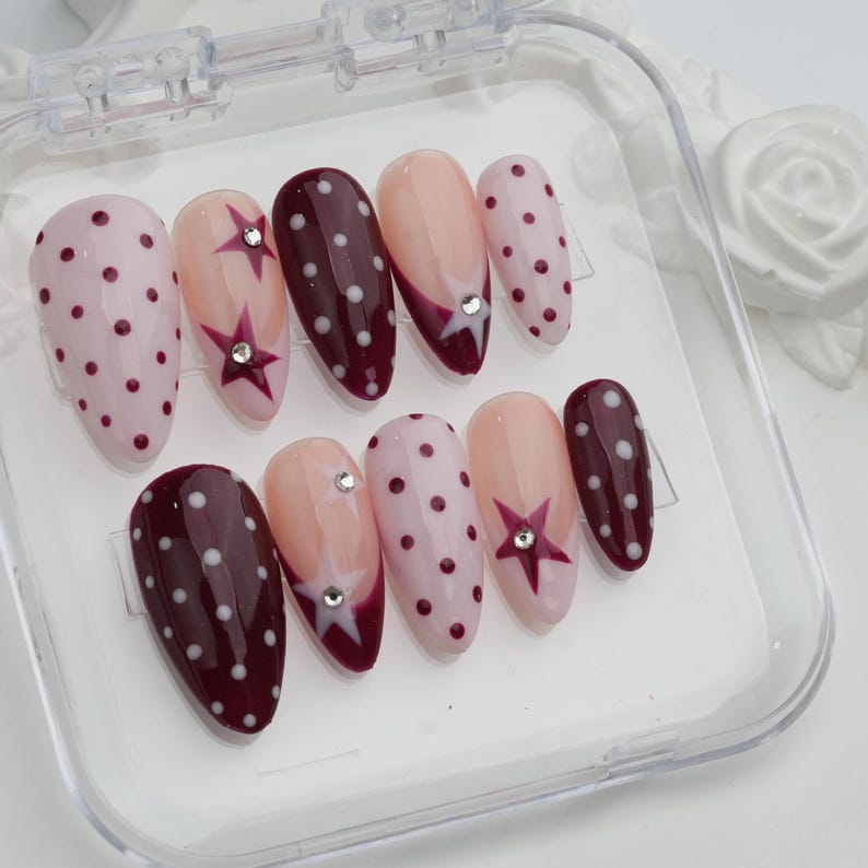May include: A set of ten artificial nails in a clear plastic case. The nails feature various designs, including polka dots, star accents, and a two-tone design with a rhinestone. Colors include pink, burgundy, and nude.
