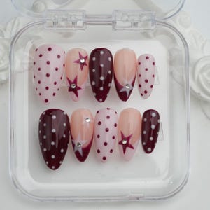 May include: A set of ten artificial nails in a clear plastic case. The nails feature various designs, including pink with white polka dots, burgundy with white polka dots, and nude with star and rhinestone accents.