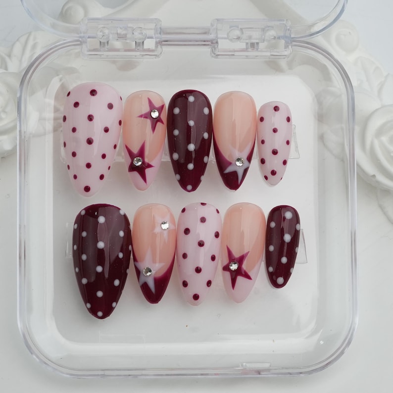 May include: A set of ten artificial nails in a clear plastic case. The nails feature various designs, including pink with white polka dots, burgundy with white polka dots, and nude with star and rhinestone accents.