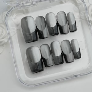 May include: A set of press-on nails in a clear plastic case. The nails are oval-shaped with a gradient design, transitioning from light gray to dark gray. The tips are a solid dark gray color.
