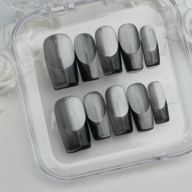May include: A set of press-on nails in a clear plastic case. The nails are oval-shaped with a gradient design, transitioning from light gray to dark gray. The tips are a solid dark gray color.
