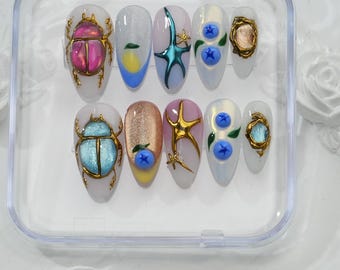 May include: A set of ten artificial nails in a clear plastic case. The nails feature various designs, including iridescent beetles, gold starfish, and blue blueberries. The nails are oval-shaped and have a glossy finish.