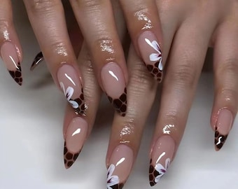 Può includere: Primo piano di una mano con unghie lunghe e a forma di mandorla. Le unghie sono dipinte di un colore nude chiaro e hanno un design floreale marrone e bianco sulle punte. Il design presenta un motivo a nido d'ape e fiori bianchi con centri bordeaux.