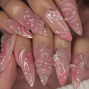 Puede incluir: Primer plano de uñas con arte de uñas rosa y blanco con flores y mariposas 3D. Las uñas son largas y puntiagudas, y las flores están hechas de plástico transparente con detalles en rosa y blanco. Las mariposas también están hechas de plástico transparente con detalles en rosa y blanco.