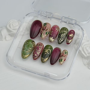 May include: A set of ten artificial nails in a clear plastic case. The nails feature a variety of designs, including solid burgundy, floral patterns, and gold accents. The colour palette includes burgundy, green, pink, and gold.