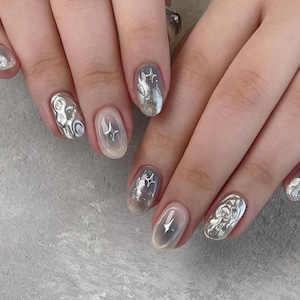 May include: Close-up of a hand with silver glitter nail polish and various silver nail art designs. The designs include stars, swirls, and abstract shapes.