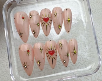 Pode incluir: Um conjunto de dez unhas postiças cor-de-rosa com glitter dourado e detalhes de coração vermelho. As unhas são exibidas em uma caixa de plástico transparente.