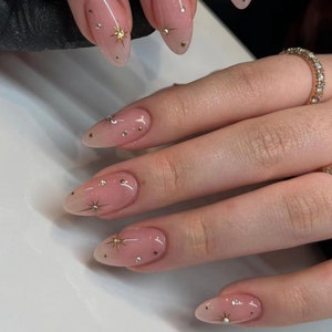 May include: Close-up of a hand with almond-shaped nails painted with a sheer pink polish and adorned with small gold stars and rhinestones.