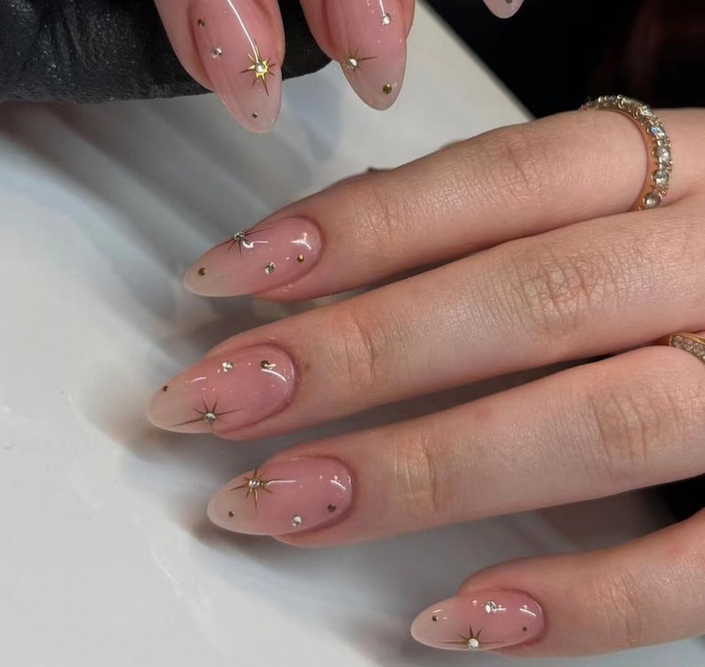 May include: Close-up of a hand with almond-shaped nails painted with a sheer pink polish and adorned with small gold stars and rhinestones.
