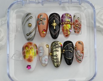 May include: A set of ten artificial nails in a clear plastic case. The nails feature various designs, including gold accents, star-shaped embellishments, and abstract patterns in colors like black, orange, and white.