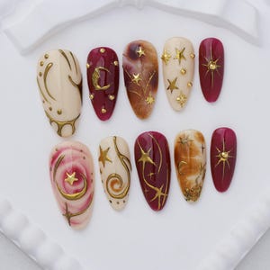 May include: A set of ten almond-shaped press-on nails featuring a celestial theme. The designs include deep burgundy, cream, and marbled brown bases adorned with metallic gold stars, crescent moons, and small raised gold studs and accents.