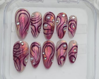 May include: A set of ten oval-shaped press-on nails. Each nail features a swirling design in shades of pink and purple, accented with silver rhinestones and small pearl-like embellishments. The nails are displayed in a clear plastic case.