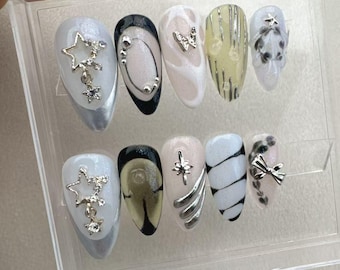 Pode incluir: Um conjunto de dez unhas postiças com diferentes designs. Os designs incluem estrelas, redemoinhos, listras e laços. As unhas são todas brancas com detalhes prateados.