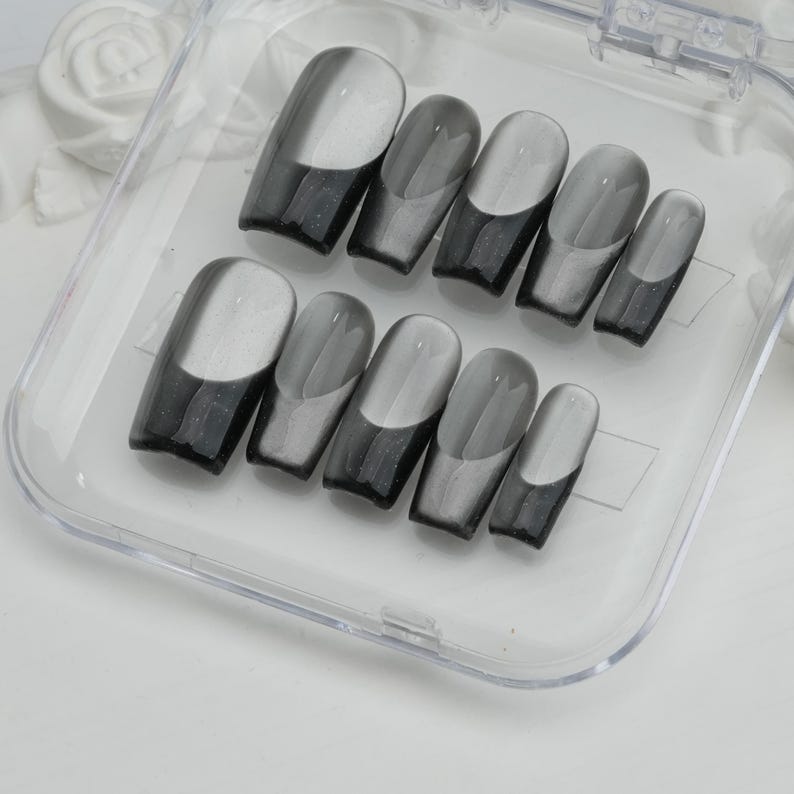 May include: A set of ten gray and black press-on nails in a clear plastic case. The nails have a glossy finish with a gradient design, transitioning from black at the base to gray at the tip. The nails are in various sizes.