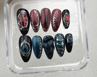 May include: A set of ten artificial nails in a clear plastic case. The nails feature various designs, including red and teal eye motifs, and dark red and teal patterns with silver accents. The nails are oval-shaped.