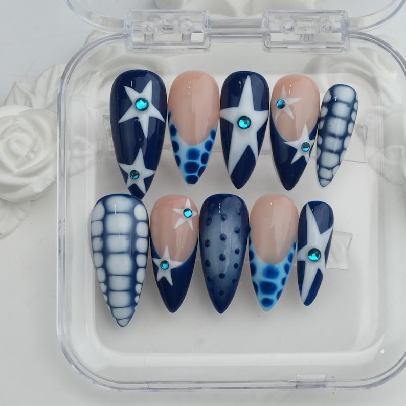 May include: A set of ten artificial nails in a clear plastic case. The nails feature a variety of designs, including navy blue with white stars, light blue and white patterns, and nude-colored nail beds with blue accents. Each nail has a small, round, blue gem.