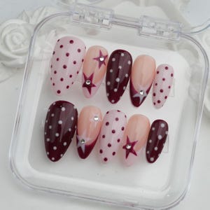 May include: A set of ten artificial nails in a clear plastic case. The nails feature various designs, including polka dots, star accents, and a French tip design. Colors include pink, burgundy, and nude.