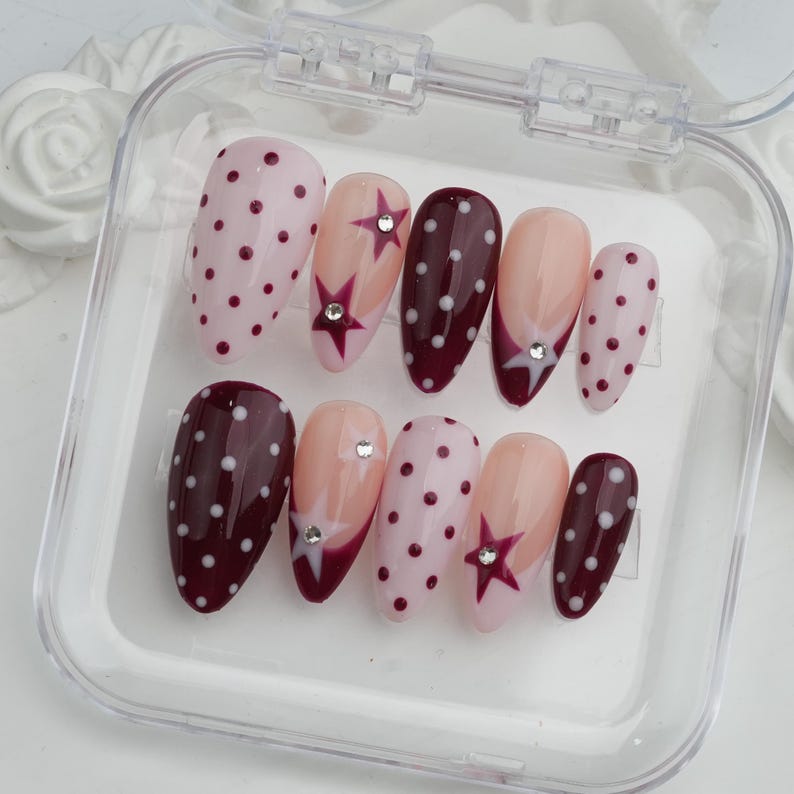May include: A set of ten artificial nails in a clear plastic case. The nails feature various designs, including polka dots, star accents, and a French tip design. Colors include pink, burgundy, and nude.