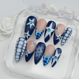 May include: A set of ten artificial nails in a clear plastic case. The nails feature a variety of designs, including navy blue with white star patterns, light pink with blue accents, and blue and white grid patterns. Each nail has a small, round, blue gem.