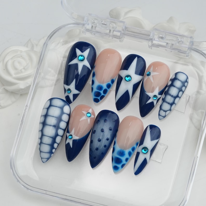 May include: A set of ten artificial nails in a clear plastic case. The nails feature a variety of designs, including navy blue with white star patterns, light pink with blue accents, and blue and white grid patterns. Each nail has a small, round, blue gem.