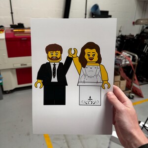K&ouml;nnte beinhalten: Eine wei&szlig;e Karte mit einer Zeichnung eines Lego-Brautpaares. Die Braut tr&auml;gt ein wei&szlig;es Kleid mit einem glitzernden Oberteil und einen wei&szlig;en Schleier. Der Br&auml;utigam tr&auml;gt einen schwarzen Anzug mit einer schwarzen Krawatte und einem wei&szlig;en Hemd. Beide l&auml;cheln und halten sich an den H&auml;nden.