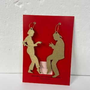 May include: Two gold wooden ornaments shaped like silhouettes of people dancing. The ornaments are hanging from red cardstock with the text "www.etsy.com/shop/waffleteeth @waffleteeth".