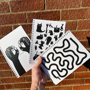 Op de afbeelding: Drie spiraalgebonden notitieboekjes met zwart-wit illustraties. Een notitieboekje toont een zwart-wit illustratie van een persoon met zijn handen die zijn ogen bedekken. Een ander notitieboekje toont een zwart-wit illustratie van een doolhof. Het derde notitieboekje toont een zwart-wit illustratie van katten.
