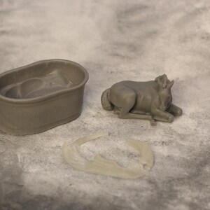 May include: A gray miniature bathtub, a reclining bull figurine, and a crescent-shaped object are arranged on a textured surface. The bull is lying down with its head raised. The bathtub is oval-shaped.
