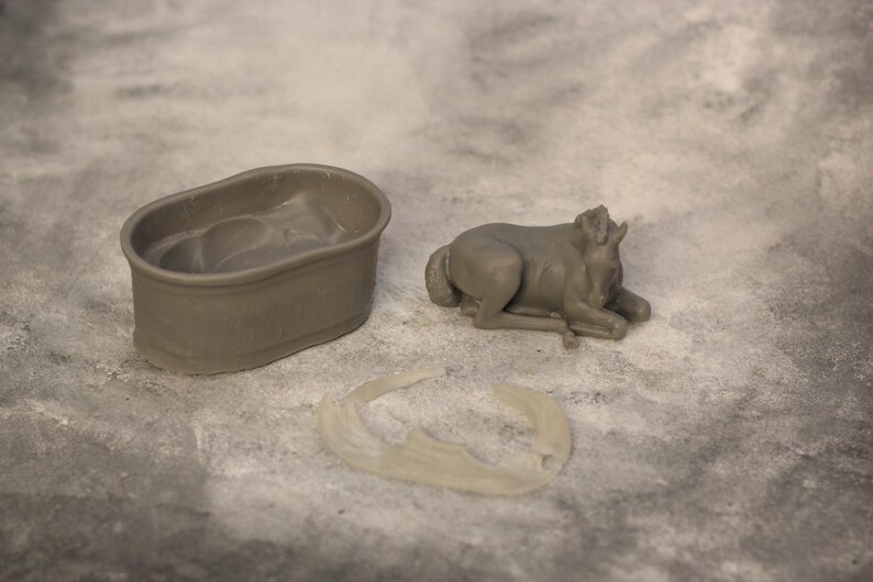 May include: A gray miniature bathtub, a reclining bull figurine, and a crescent-shaped object are arranged on a textured surface. The bull is lying down with its head raised. The bathtub is oval-shaped.