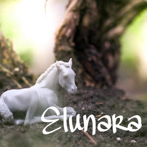 May include: A white figurine of a horse lying down on a bed of soil. The horse has a detailed mane and tail. The word "Elunara" is written in a cursive font below the horse.