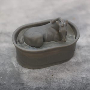 May include: A gray figurine of a horse resting in a bathtub. The horse is lying down with its head resting on its front legs. The bathtub is oval-shaped and has a simple design. The figurine is made of a solid material.