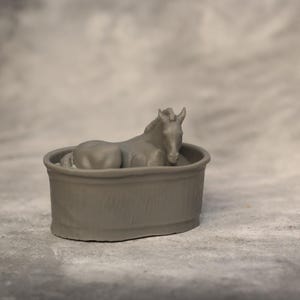 May include: A gray figurine of a horse resting inside a round, gray tub. The horse is lying down with its head and neck raised. The tub has a simple, rounded design. The background is a mottled gray.
