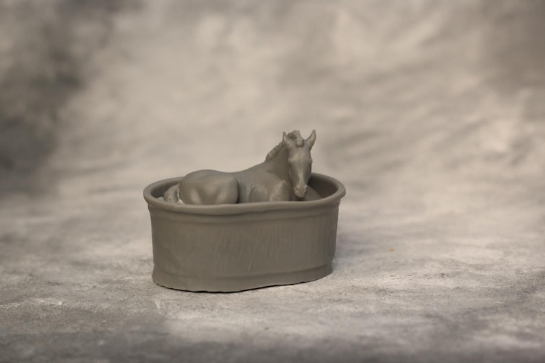 May include: A gray figurine of a horse resting inside a round, gray tub. The horse is lying down with its head and neck raised. The tub has a simple, rounded design. The background is a mottled gray.