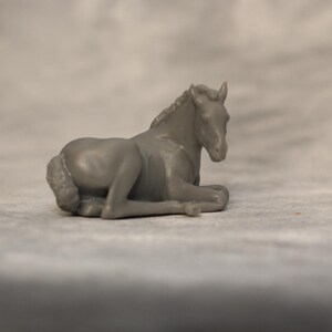 May include: A gray figurine of a resting horse. The horse is lying down with its legs tucked beneath its body. The figurine is detailed, showing the horse's mane, tail, and facial features. The background is a blurred, neutral tone.