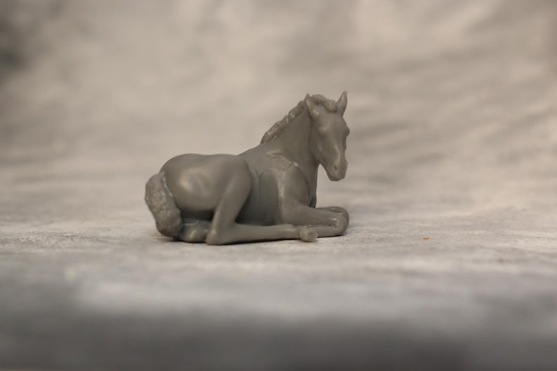 May include: A gray figurine of a resting horse. The horse is lying down with its legs tucked beneath its body. The figurine is detailed, showing the horse's mane, tail, and facial features. The background is a blurred, neutral tone.