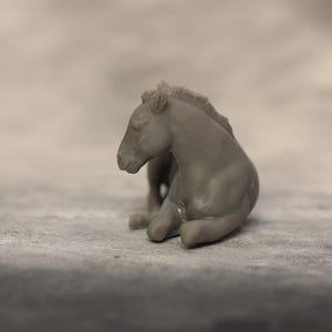 May include: A small, gray figurine of a resting horse. The horse is in a seated position, with its legs tucked beneath its body. The figurine has a smooth texture and is set against a neutral background.