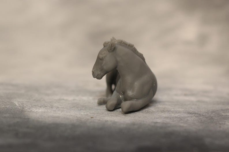 May include: A small, gray figurine of a resting horse. The horse is in a seated position, with its legs tucked beneath its body. The figurine has a smooth texture and is set against a neutral background.