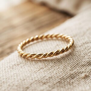 Op de afbeelding: Een dunne, ronde ring gemaakt van gepolijst goudkleurig metaal, voorzien van een doorlopende gedraaide touwtextuur die een herhalend spiraalpatroon rond de gehele omtrek van de band vormt, met een heldere, reflecterende metalen afwerking.