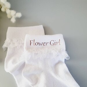 Puede incluir: Calcetines blancos hasta el tobillo con ribete de encaje y las palabras "Flower Girl" bordadas en la parte superior.
