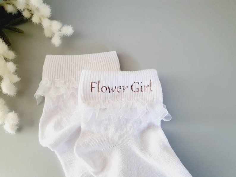 Puede incluir: Calcetines blancos hasta el tobillo con ribete de encaje y las palabras "Flower Girl" bordadas en la parte superior.